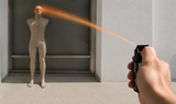 A hand holds a black spray bottle and aims the orange-colored spray toward a mannequin torso in front of a window.